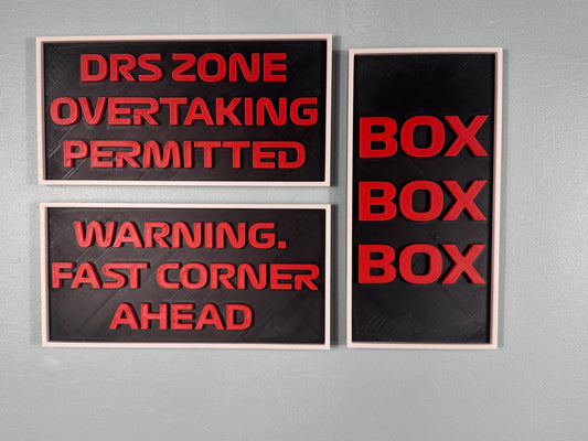 3 wall decor signs, in the f1 font and colours, black background, red writing and a white frame, measuring 250mmx125mm, 1 sign says "drs zone overtaking permitted" this sign is horizontal, another sign says "warning. fast corner ahead" this sign is also horizontal and the 3rd verticle sign says "box box box "